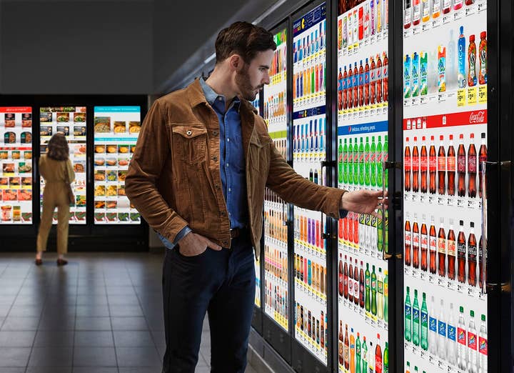 A man in jeans, a blue denim shirt, and a brown denim jacket looks at cooler with a TV monitor instead of a glass door, and reaches for the handle