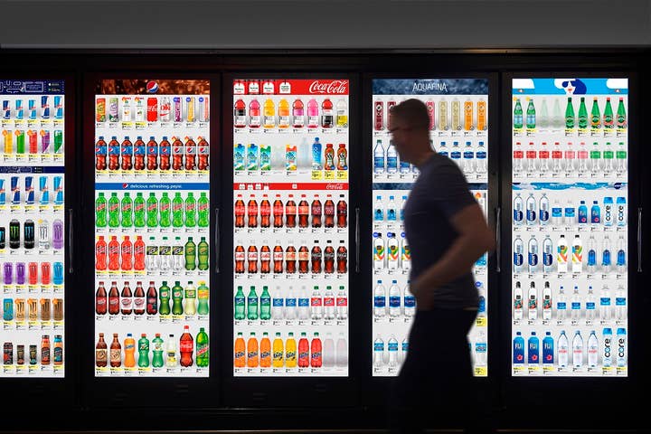 A bank of grocery store coolers with blindingly bright screens instead of glass doors show pictures of soda, water, and juice bottles. A dark and blurry figure that appears to be a man walks by, seemingly looking at the screens as he passes